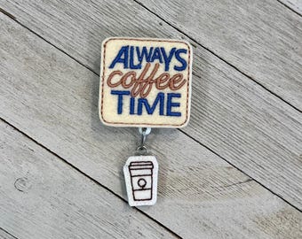 May include: A cream-colored badge reel with the text "ALWAYS coffee TIME" in blue and brown embroidery. A small white coffee cup charm hangs below. The reel is on a light wood-grain surface.