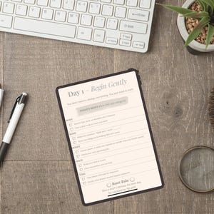 Puede incluir: Una vista cenital de un escritorio de madera con una tableta que muestra una lista de verificación diaria titulada "Day 1 - Begin Gently". Un teclado blanco, un bloc de notas, un bolígrafo, una planta, una taza de café y una lupa también están en el escritorio.