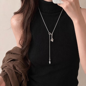 May include: A silver-toned lariat necklace featuring a delicate snake chain, a small calla lily flower pendant, and a slender leaf-shaped charm at the end of the drop. The necklace is worn over a black sleeveless ribbed turtleneck top.