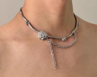 May include: A silver necklace with a floral pendant and chain details. The necklace has a curved design with decorative elements and a dangling chain. The jewellery is worn around the neck.