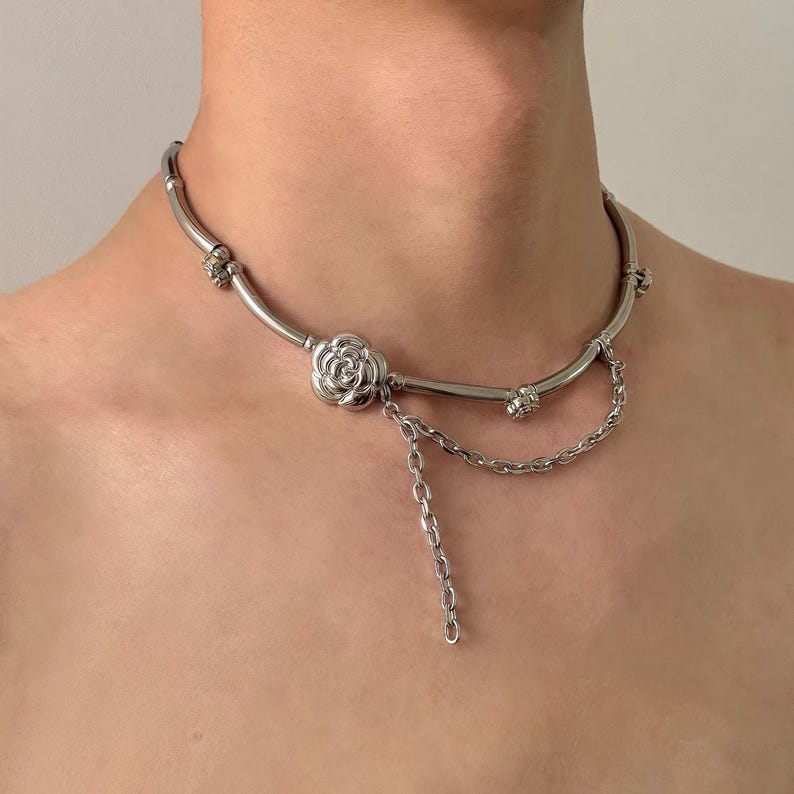K&ouml;nnte beinhalten: Eine silberfarbene Halskette mit einem floralen Anh&auml;nger und Kettenakzenten. Die Halskette hat ein geschwungenes Design mit dekorativen Elementen und einer herabh&auml;ngenden Kette. Der Schmuck wird um den Hals getragen.