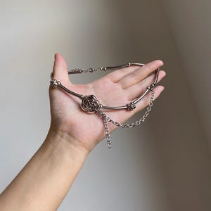 K&ouml;nnte beinhalten: Silberfarbene Halskette mit einem zentralen Rosenanh&auml;nger und zarten Kettenakzenten. Die Halskette hat ein flexibles Design mit einer Reihe kleiner, dekorativer Elemente entlang des Bandes. Der Schmuck wird in einer Hand vor neutralem Hintergrund gehalten.