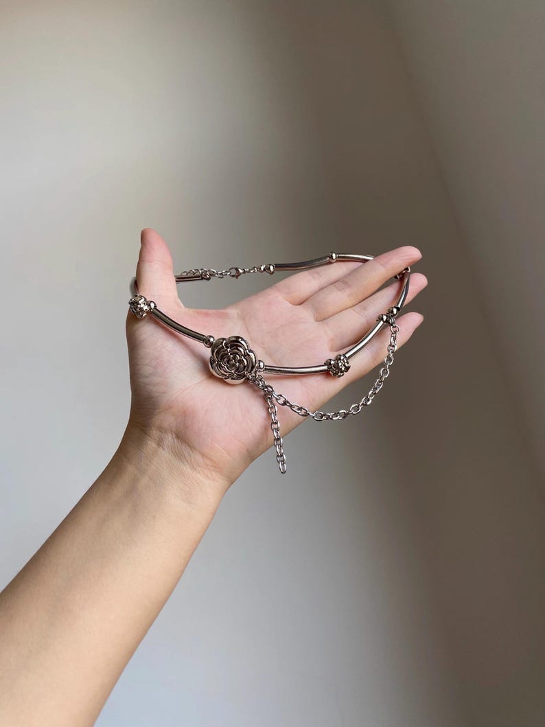 K&ouml;nnte beinhalten: Silberfarbene Halskette mit einem zentralen Rosenanh&auml;nger und zarten Kettenakzenten. Die Halskette hat ein flexibles Design mit einer Reihe kleiner, dekorativer Elemente entlang des Bandes. Der Schmuck wird in einer Hand vor neutralem Hintergrund gehalten.