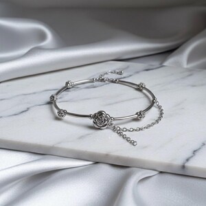 K&ouml;nnte beinhalten: Ein silbernes Armband mit floralem Design. Das Armband hat einen rosenf&ouml;rmigen Anh&auml;nger in der Mitte und kleinere florale Akzente entlang des Bandes. Eine zarte Kette h&auml;ngt von der Rose herab. Das Armband wird auf einer wei&szlig;en Marmoroberfl&auml;che mit seidigem, wei&szlig;em Hintergrund pr&auml;sentiert.