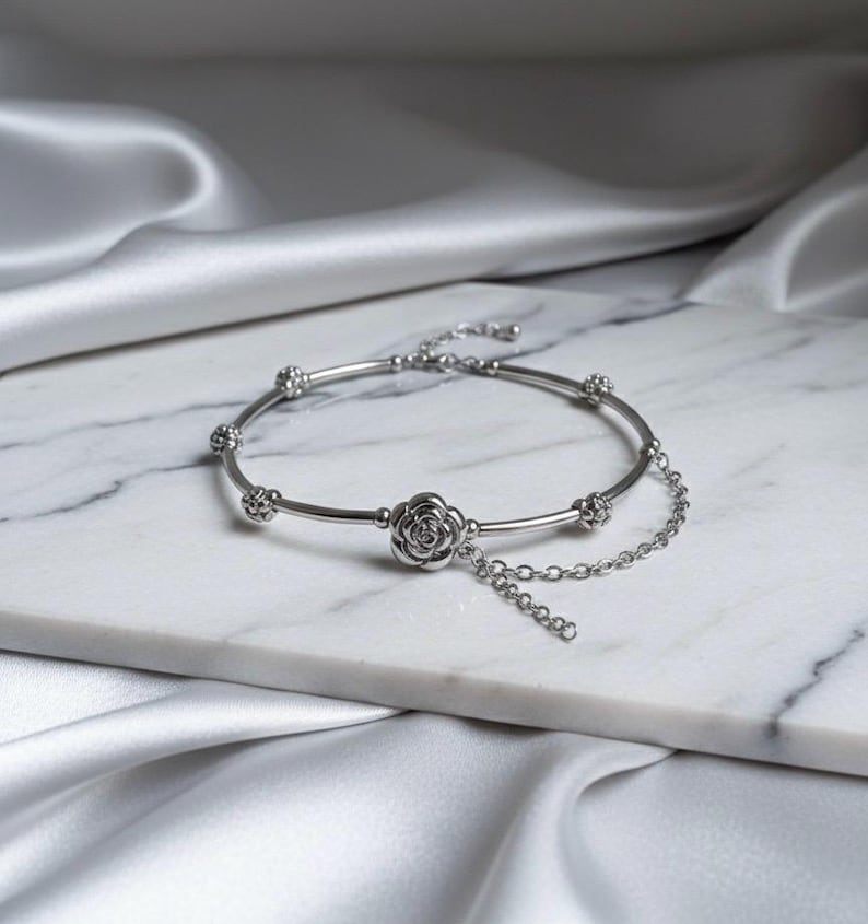 K&ouml;nnte beinhalten: Ein silbernes Armband mit floralem Design. Das Armband hat einen rosenf&ouml;rmigen Anh&auml;nger in der Mitte und kleinere florale Akzente entlang des Bandes. Eine zarte Kette h&auml;ngt von der Rose herab. Das Armband wird auf einer wei&szlig;en Marmoroberfl&auml;che mit seidigem, wei&szlig;em Hintergrund pr&auml;sentiert.