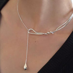 May include: A silver necklace featuring a unique design. The necklace has a curved bar with a dangling teardrop pendant. The necklace is worn on a person's neck, and the design is visible against a dark background.
