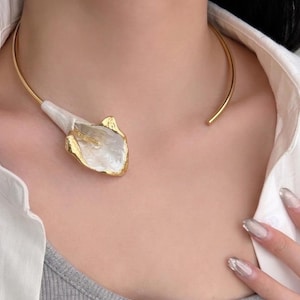 May include: A gold-tone collar necklace featuring a white and gold calla lily pendant. The pendant has a pearlescent finish with gold edging. The necklace is worn around the neck, with the open ends at the back.