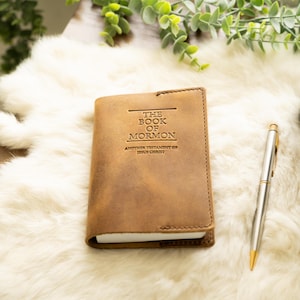 May include: A tan leather cover for The Book of Mormon, Another Testament of Jesus Christ, featuring visible stitching along the edges. The cover is positioned next to a silver metal pen, showcasing a smooth, matte texture and embossed lettering.