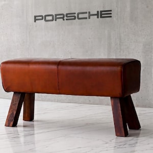 May include: A rectangular bench featuring a smooth, cognac-colored leather top with a central seam, supported by four thick, dark brown wooden legs angled outward. The bench is positioned in front of a concrete wall with the word PORSCHE in black.