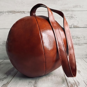 May include: A spherical handbag crafted from smooth, glossy brown material. The bag features a rounded, ball-like shape with visible stitched seams and a single, long, matching brown strap attached to the top for carrying or shoulder wear.