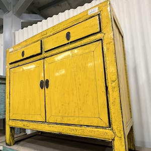 May include: A bright yellow wooden cabinet featuring two top drawers and two lower cabinet doors with dark metal hardware. The piece has a distressed finish with visible wood grain and black accents along the edges and corners of the frame.