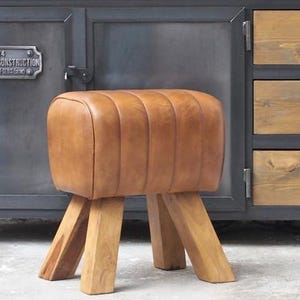 Op de afbeelding: Een bruine leren en houten kruk met een rechthoekige zitting en vier schuine houten poten. De kruk staat voor een donkere kast met houten laden en een metalen frame. De zitting van de kruk heeft een geribbeld ontwerp.