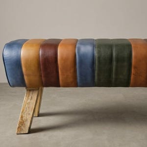 Könnte beinhalten: Eine rechteckige Bank mit einer gepolsterten Sitzfläche, die mit vertikalen Paneelen aus mehrfarbigem Leder in Blau-, Beige-, Braun- und Grüntönen bezogen ist. Die Sitzfläche ruht auf vier schrägen Holzbeinen mit heller, natürlicher Maserung.