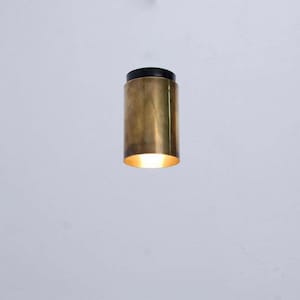 May include: A cylindrical pendant light fixture with a brass finish and a black top. The light is on, emitting a warm glow from the bottom. The fixture is hanging against a white background.