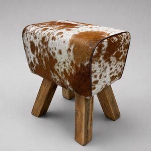 May include: A rectangular stool featuring a seat upholstered in brown and white cowhide with a smooth texture. The seat is supported by four thick, sturdy legs crafted from natural light brown wood with a visible grain and a rough, rustic finish.