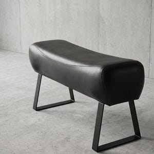 May include: A black leather bench with a smooth, curved top surface and rounded edges. The seat is supported by two black metal legs, each featuring a trapezoidal frame design that provides a stable base for the elongated, padded seating area.