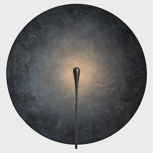 May include: A circular wall light fixture featuring a dark, mottled gray and black textured surface. A slender, vertical black stem extends from the bottom toward the center, where a soft, warm, glowing light emanates from behind the fixture.