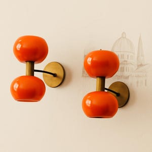 Op de afbeelding: Twee oranje wandkandelaars met een modern design uit het midden van de eeuw. Elke wandkandelaar heeft twee afgeronde oranje tinten, een messingkleurige steel en een cirkelvormige messing basis. Een zwarte draad verbindt de wandkandelaar met de muur.