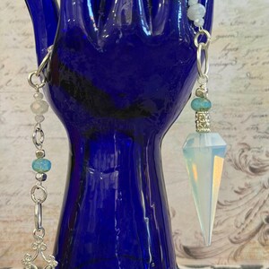 May include: A silver-toned pendulum with a faceted, opalescent crystal point and turquoise and clear bead accents. The pendulum hangs from a chain with a decorative filigree element. The background is a blue glass hand.