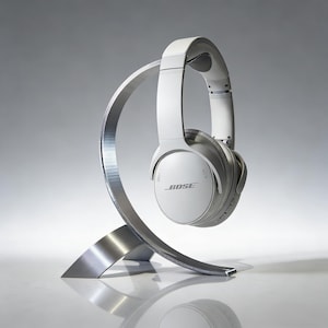 May include: White Bose headphones displayed on a silver headphone stand. The stand has a curved, modern design. The headphones are white with the Bose logo. The background is a gradient of grey. The stand is made of metal.