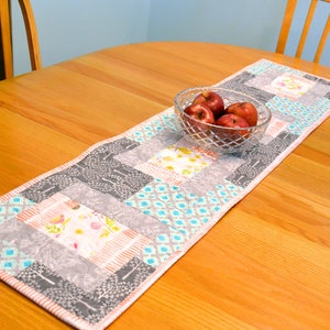 May include: A patchwork table runner with a variety of patterns in shades of gray, blue, and pink. The runner is laid out on a wooden table with a bowl of red apples in the center.
