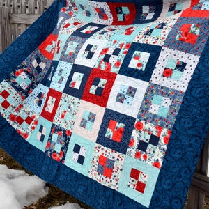 May include: A blue and white quilt with a patchwork design featuring red, white, and blue squares. The quilt is made of various fabrics with floral and geometric patterns. It is hanging on a wooden fence.