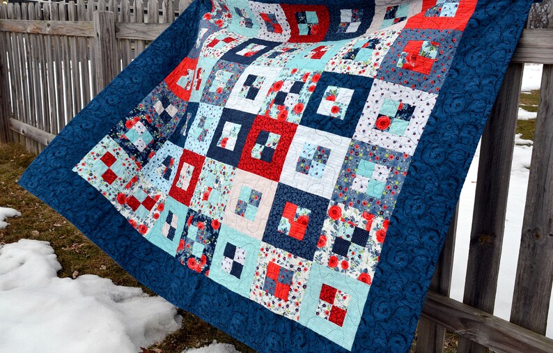May include: A blue and white quilt with a patchwork design featuring red, white, and blue squares. The quilt is made of various fabrics with floral and geometric patterns. It is hanging on a wooden fence.