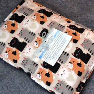 May include: A beige fabric heating pad with a cat print. The cats are in various colors, including black, gray, orange, and white. The pad has a tag attached that says "Cozy Bags".