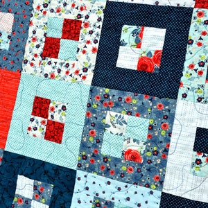 May include: A quilt made of various fabrics in shades of blue, red, and white. The quilt is made up of squares with different patterns, including floral prints, polka dots, and stripes. The quilt is quilted with a simple stitch pattern.