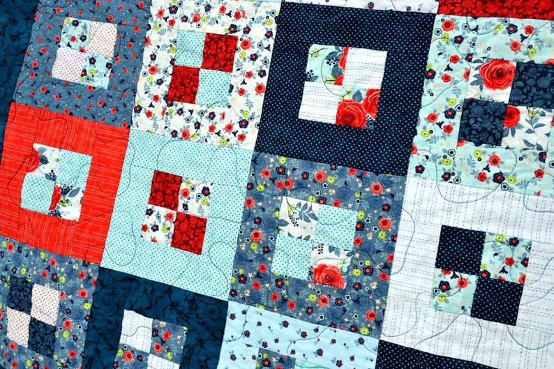 May include: A quilt made of various fabrics in shades of blue, red, and white. The quilt is made up of squares with different patterns, including floral prints, polka dots, and stripes. The quilt is quilted with a simple stitch pattern.