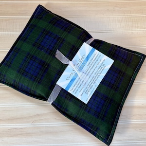 May include: A rectangular heating pad featuring a dark green and navy blue plaid pattern. A white ribbon is tied around the center, securing a white instructional card with blue text that reads Cozy Corn Bags and includes usage instructions.