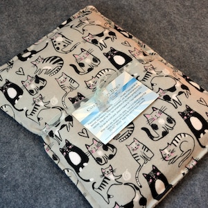 May include: A gray fabric heating pad with a black and white cat print. The pad has a white tag with the text "Cozy in Paws" and instructions for use.