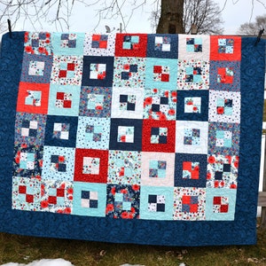 May include: A quilt made of blue, red, and white fabric with a floral pattern. The quilt is hanging on a wooden fence.
