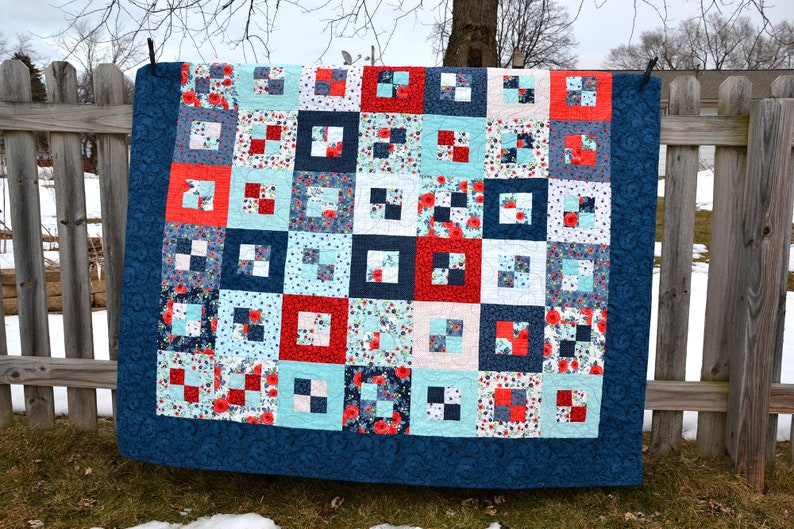 May include: A quilt made of blue, red, and white fabric with a floral pattern. The quilt is hanging on a wooden fence.