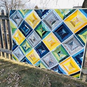May include: A quilt with a geometric pattern in shades of blue, green, yellow, and gray. The quilt is made of fabric scraps sewn together in a diamond pattern.