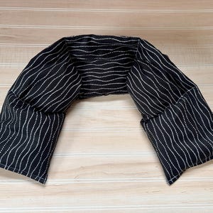 May include: A U-shaped neck heating pad made of black fabric featuring a repeating pattern of thin, white, wavy dotted lines. The fabric is segmented into multiple sections with visible stitching, designed to drape comfortably over the shoulders.