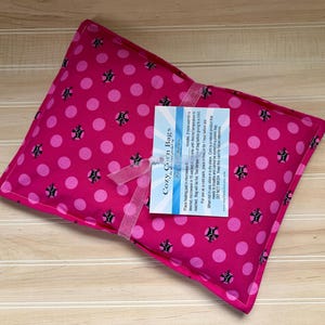 May include: A hot pink, square-shaped Cozy Corn Bags heating pad with a polka dot and floral pattern. A white tag with blue text is attached with a pink ribbon. The heating pad is on a light brown wooden surface.