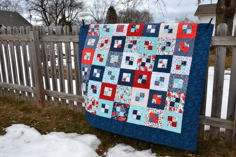May include: A blue and white quilt with a red and white floral pattern. The quilt is hanging on a wooden fence.