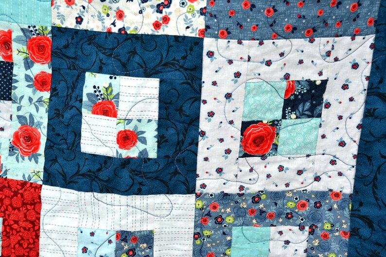 May include: A quilt made of various fabrics with floral patterns in shades of blue, white, and red. The quilt is stitched together in a patchwork design with a square pattern.