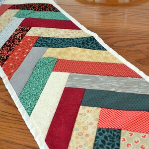 May include: A white-trimmed table runner with a chevron pattern in shades of red, green, cream, and gray.