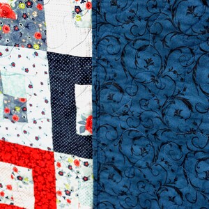 May include: A patchwork quilt with a red, white, and blue color scheme. The quilt is made of different fabrics with floral patterns and polka dots. The back of the quilt is a solid blue fabric with a black floral pattern.