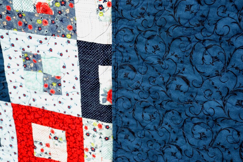 May include: A patchwork quilt with a red, white, and blue color scheme. The quilt is made of different fabrics with floral patterns and polka dots. The back of the quilt is a solid blue fabric with a black floral pattern.