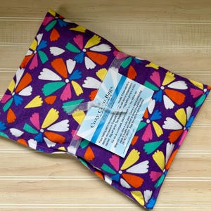 May include: A rectangular fabric heating pad featuring a purple background with a vibrant floral pattern in white, yellow, orange, pink, and teal. A white ribbon wraps around the center, holding a rectangular instruction card labeled Cozy Corn Bags.