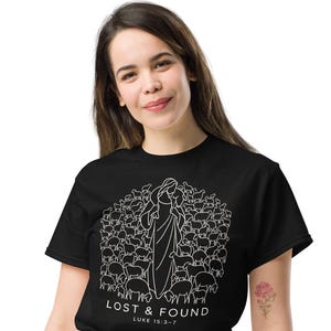 May include: A black short-sleeved t-shirt featuring a white line art graphic of a shepherd carrying a sheep surrounded by a flock. Below the graphic, the text reads "LOST & FOUND" followed by "LUKE 15:3-7" in a clean, sans-serif font.