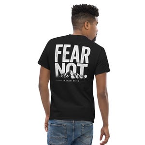 May include: Black t-shirt featuring the words "FEAR NOT" in large white letters on the back. A mountain range graphic and the text "ISAIAH 41:10" are below the text. The shirt is made of a soft material.