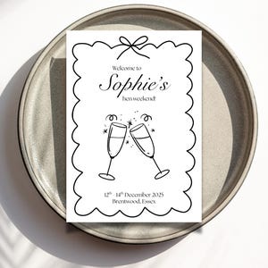 May include: A white invitation card with black text and illustrations, featuring the words "Welcome to Sophie's hen weekend!" and the date "12th - 14th December 2025". Two champagne glasses are clinking. The card is on a gray plate.