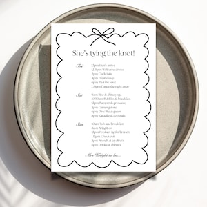 May include: A white wedding itinerary with black text, titled "She's tying the knot!" on a scalloped border, rests on a gray ceramic plate. The schedule includes times for welcome drinks, cocktails, and dancing.