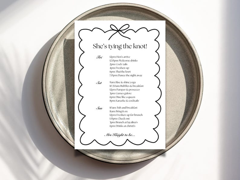 May include: A white wedding itinerary with black text, titled "She's tying the knot!" on a scalloped border, rests on a gray ceramic plate. The schedule includes times for welcome drinks, cocktails, and dancing.