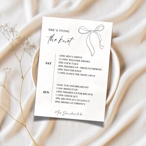 Może przedstawiać: Biała karta z napisem "She's Tying the Knot" i harmonogramem weekendu. Karta leży na białym talerzu, z suszonymi kwiatami na pierwszym planie. Harmonogram zawiera godziny i aktywności.