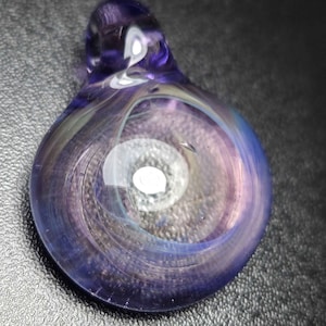 May include: A handcrafted glass pendant with a swirling design. The pendant features shades of purple, blue, and white, creating a mesmerizing, ethereal effect. The glass is smooth and rounded, with a loop at the top for a chain.